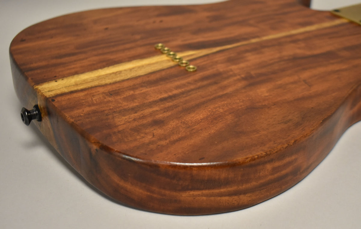 The back of a natural finish T-style guitar body, showing a light wood stripe, six brass string ferrules, and a black output jack.