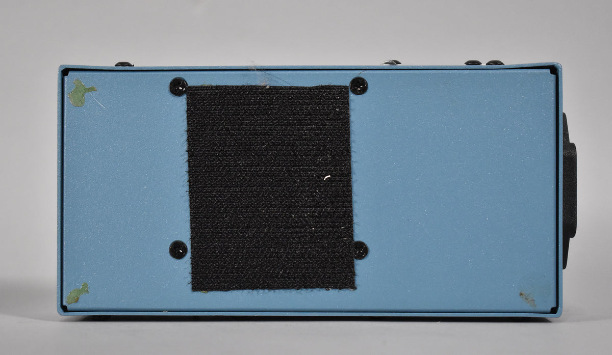 The light blue pedal power supply is seen from the bottom, with a large black velcro strip covering the center.