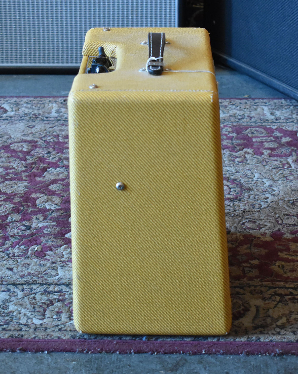 The tweed combo amp stands upright, showing its right side and top with a dark brown leather handle.