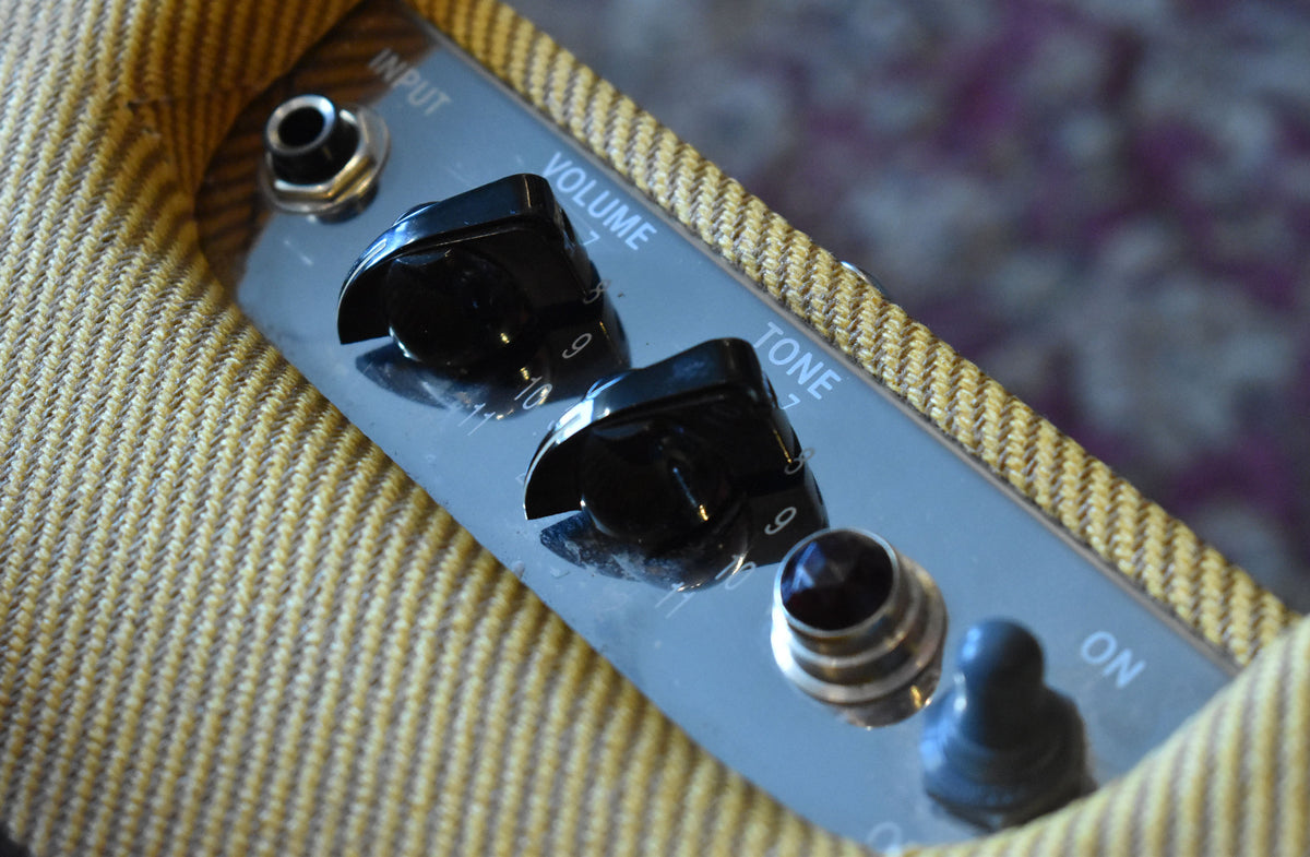 Close-up of the lacquered tweed amplifier's control panel, showing the input jack, black Volume and Tone knobs, and a black power switch.