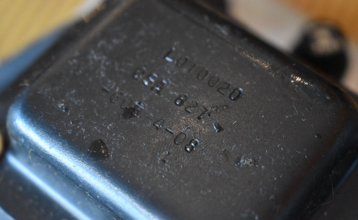Close-up of a dark grey metal component, possibly a transformer, with stamped numbers and visible wear and dust.