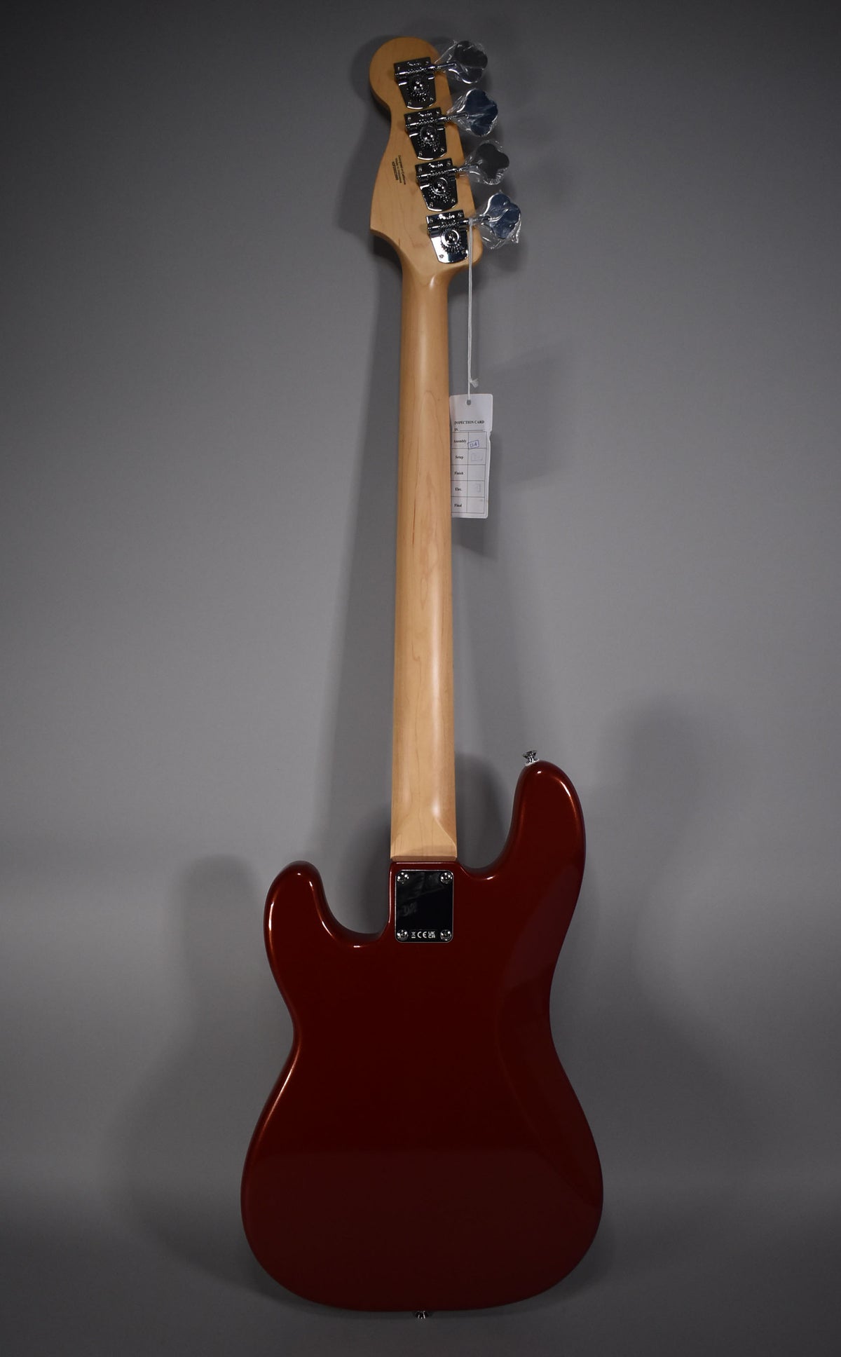 The bass is viewed from the back, showing the Candy Cola body, light maple neck, and chrome tuners with plastic wrapping.