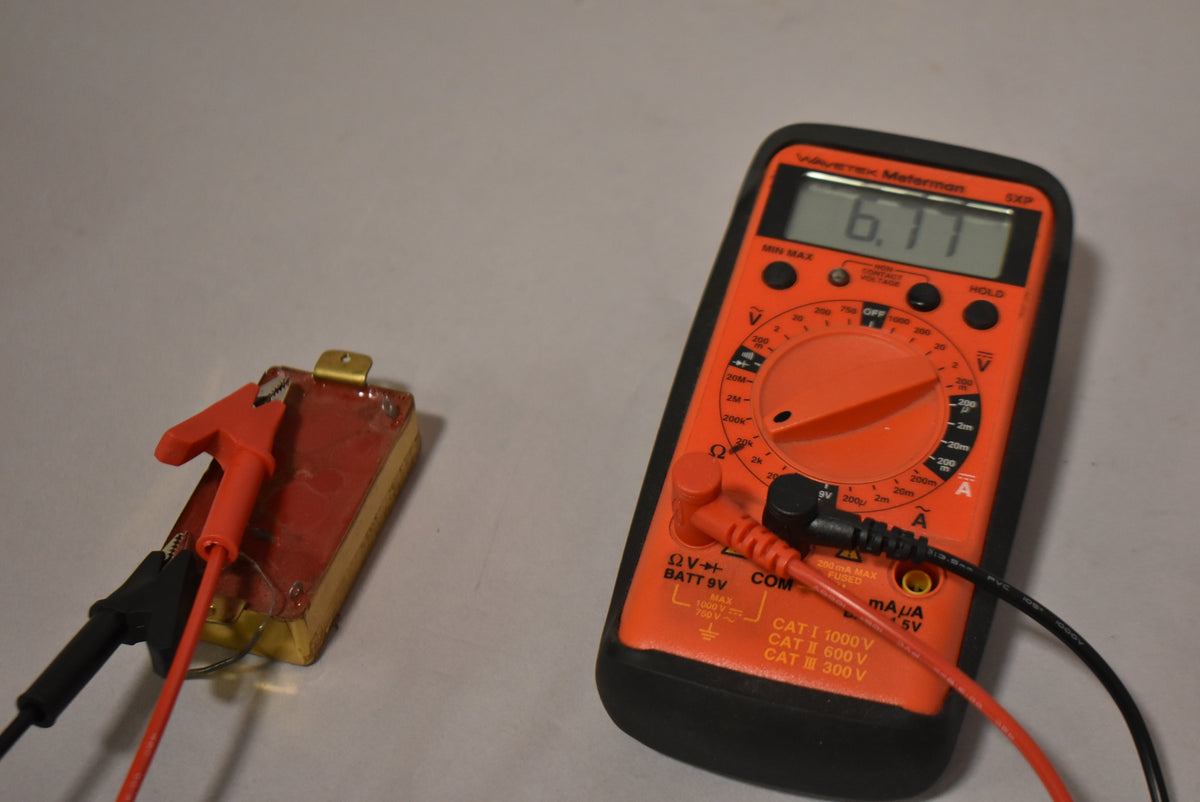 The gold humbucker pickup, with a red alligator clip and black test lead attached, is next to an orange multimeter reading 6.11.