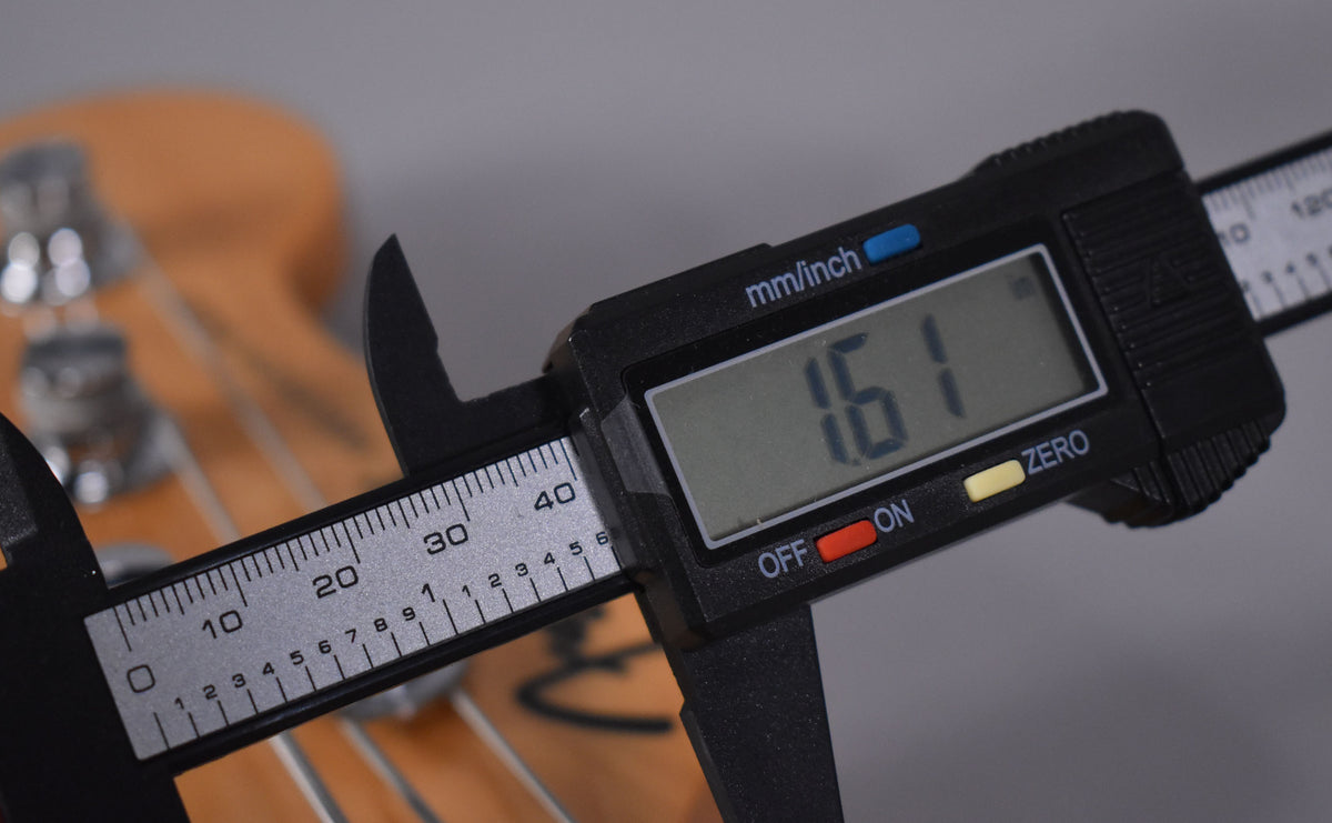 A digital caliper measures the bass's nut width at 1.61 inches, with the headstock and tuning pegs blurred in the background.
