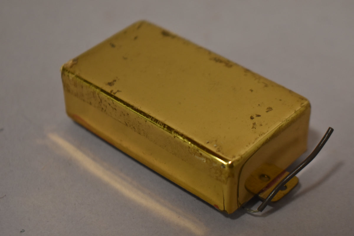 A gold humbucker pickup cover, viewed from above, shows significant wear and tarnishing on its top and sides, with two wires emerging.