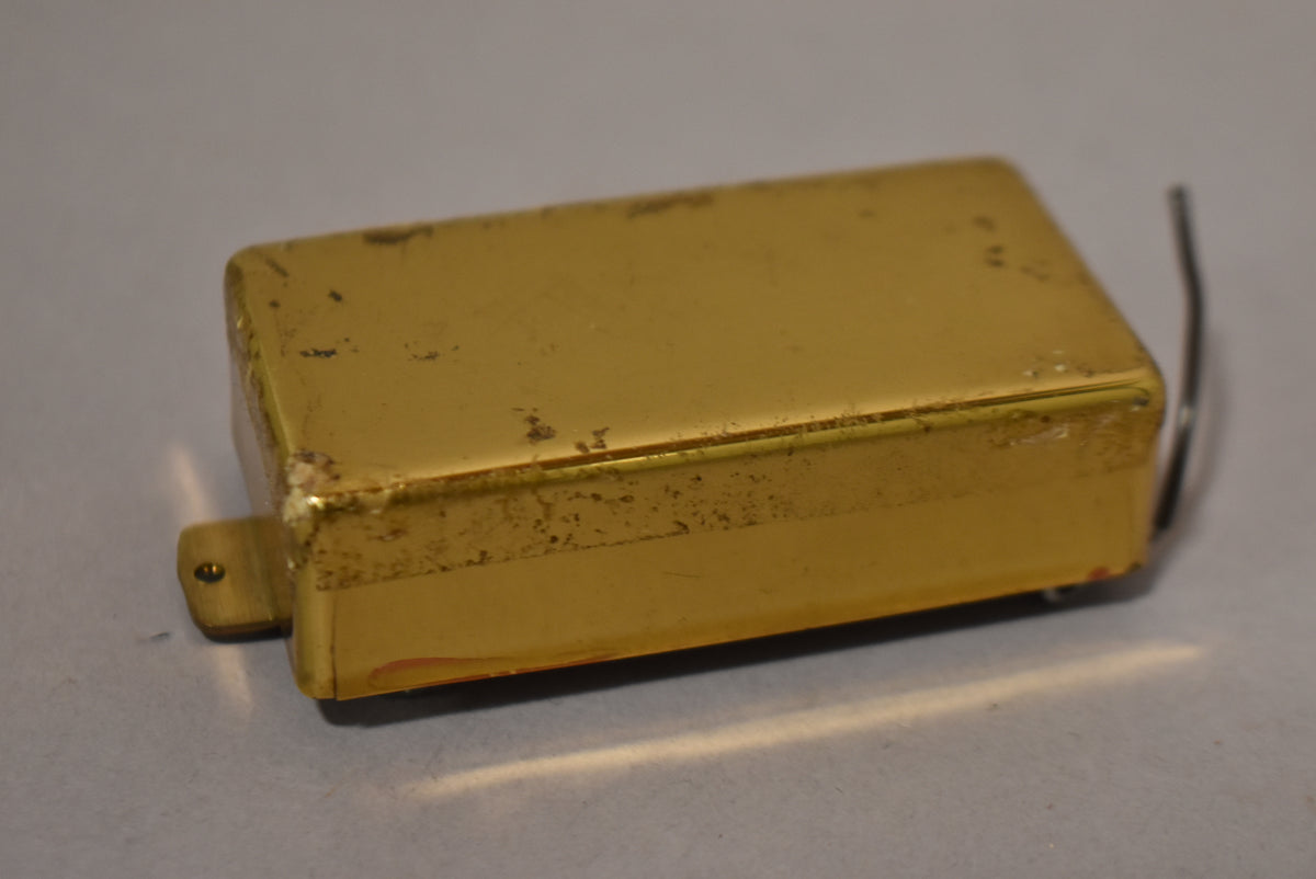 A gold humbucker pickup cover, viewed from above and slightly to the side, shows heavy wear and tarnishing, with a mounting tab visible.