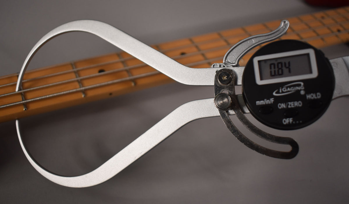 A digital caliper measures the bass's maple fretboard and strings, displaying "0.84" on its screen.