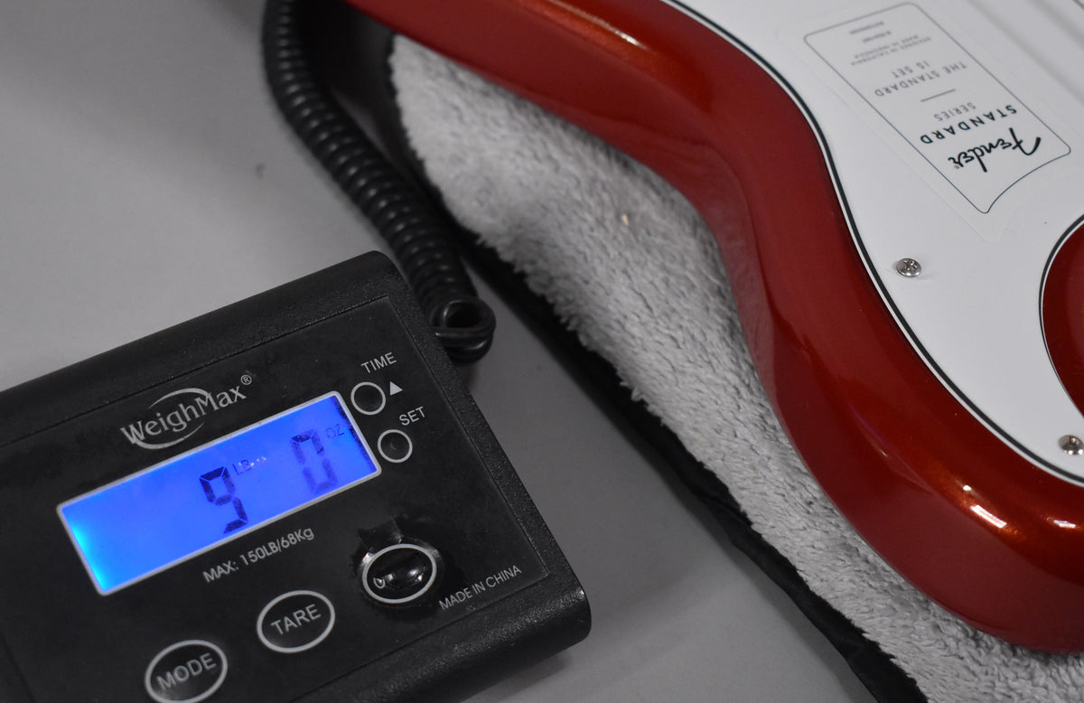 The Candy Cola bass body with a white pickguard rests on a grey towel, next to a digital scale displaying "9 Lb 0 Oz".