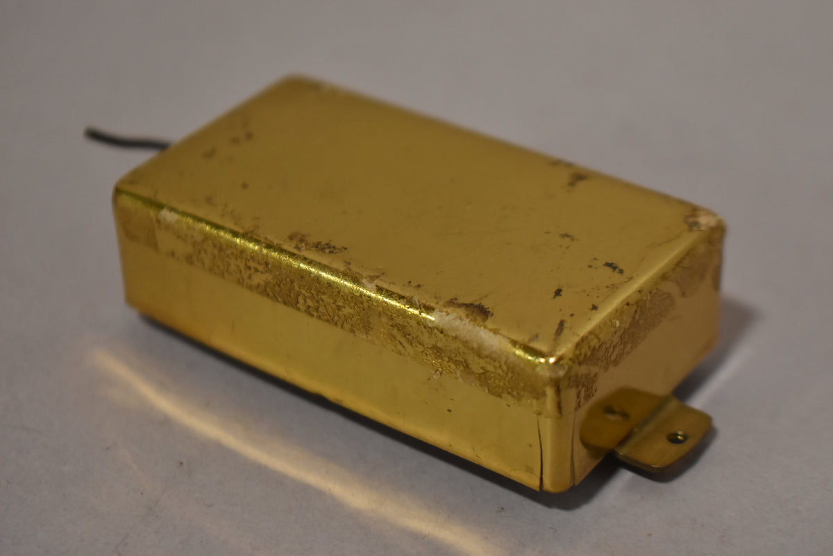A gold humbucker pickup cover, viewed from above and slightly to the side, shows significant wear, tarnishing, and flaking on its surface.