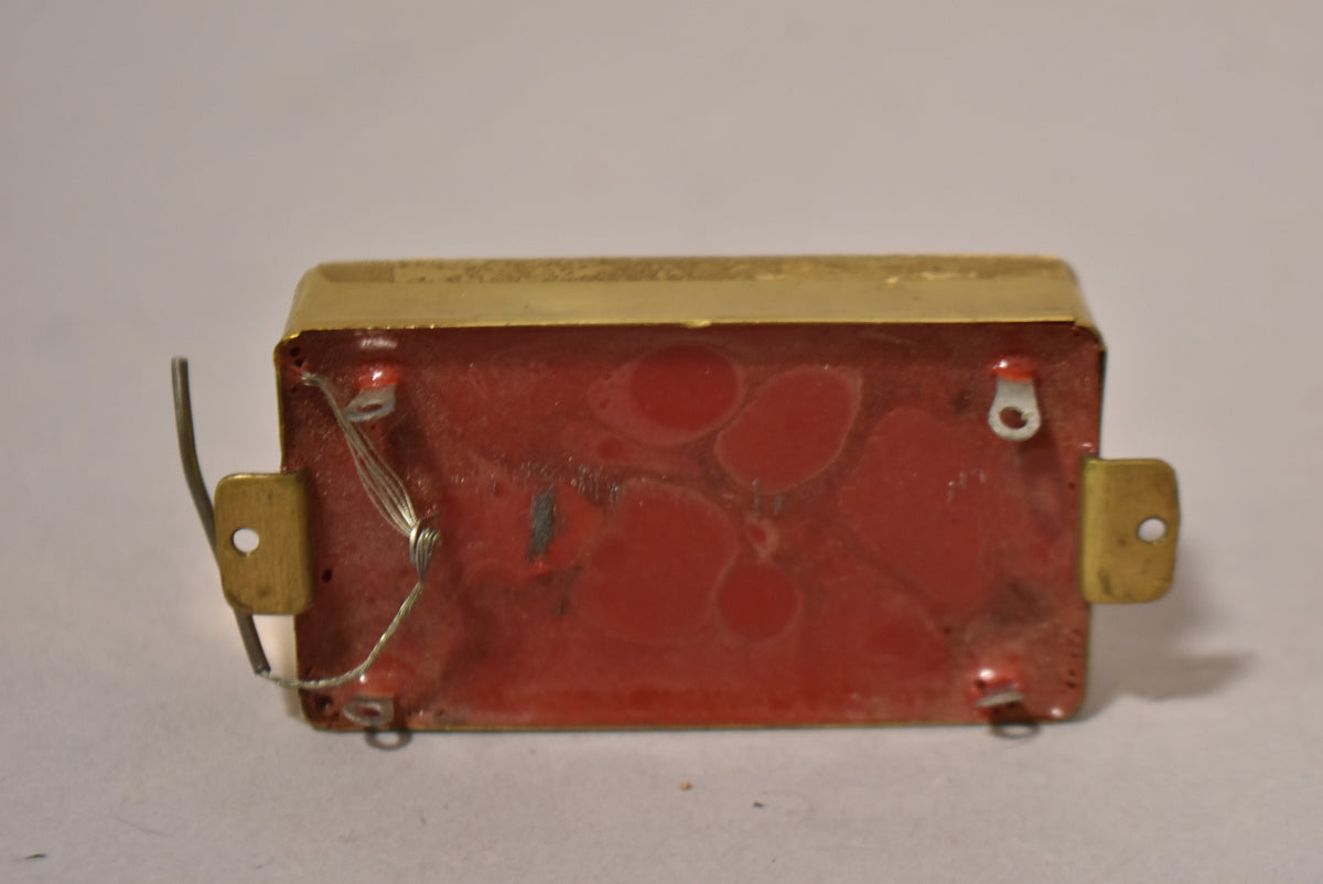 The humbucker pickup is viewed from the bottom, showing the red wax potting, gold mounting tabs, and a silver wire.