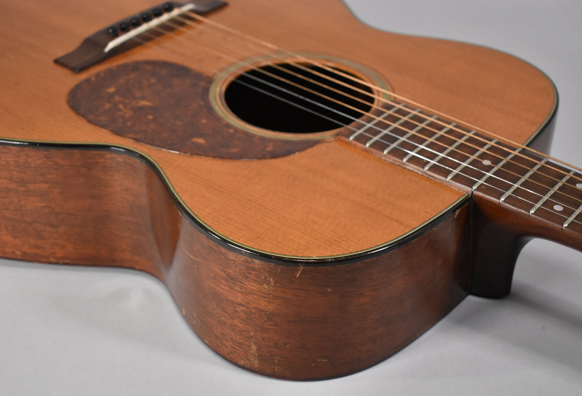 The natural finish acoustic guitar's lower bout and soundhole are visible, showing a worn pickguard and body scratches.
