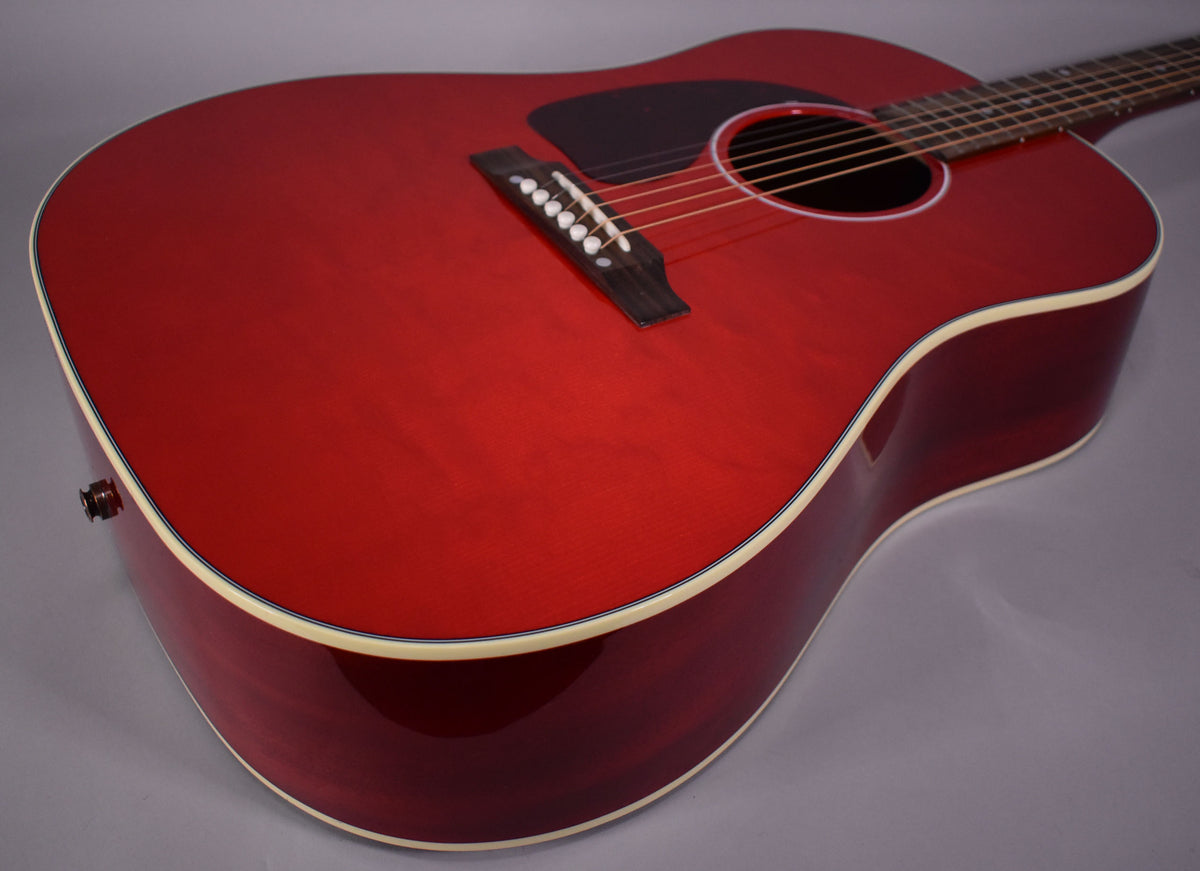 The cherry red acoustic guitar is angled from the lower bout, showing the glossy finish, black pickguard, and white binding.