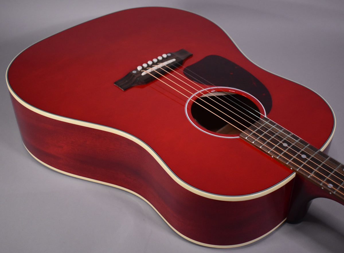 The cherry red acoustic guitar, with a dark pickguard and cream binding, is angled to show the body and part of the fretboard.