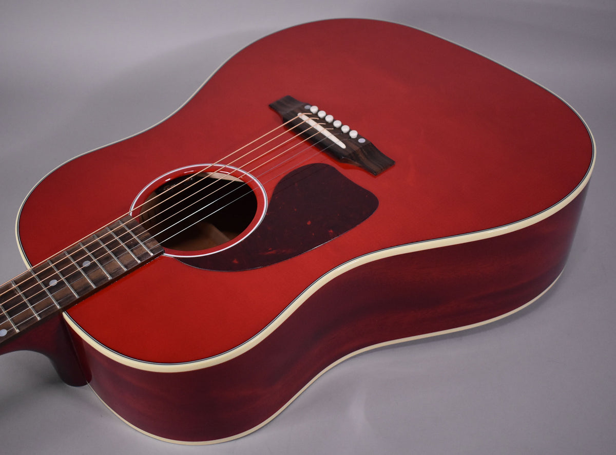 Close-up of a cherry red acoustic guitar, showing the body, soundhole, pickguard, bridge, and lower fretboard from an angle.