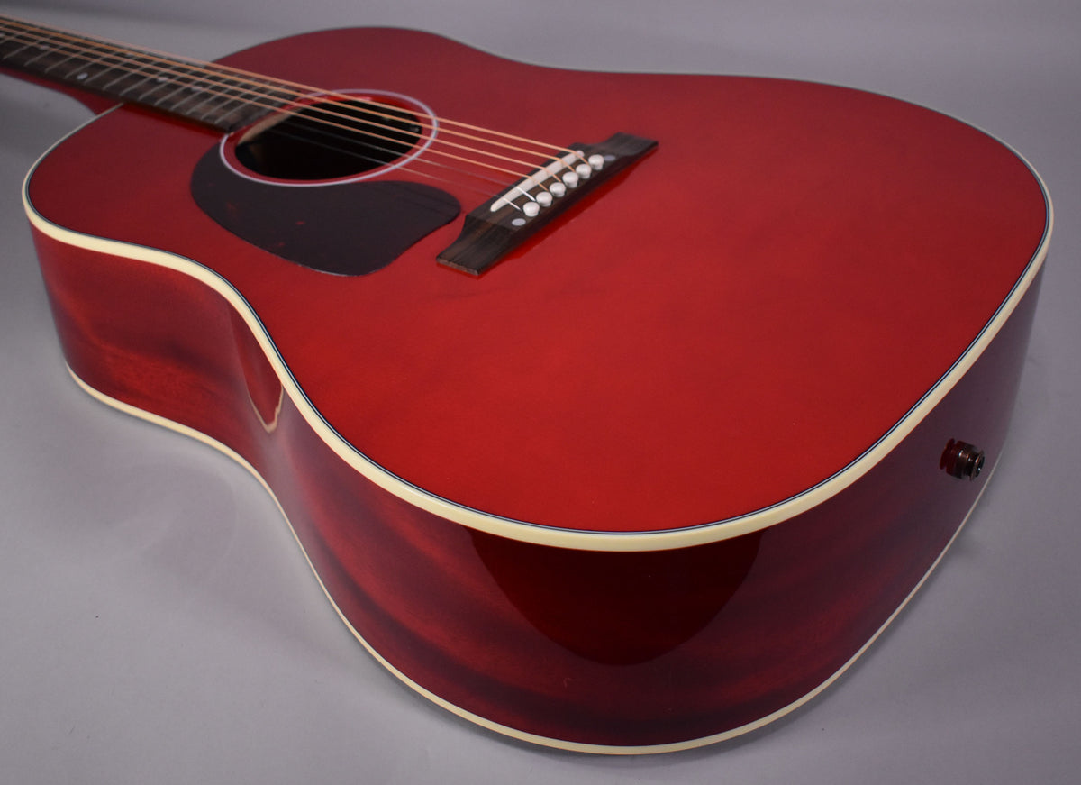 The left-handed cherry acoustic guitar's body is angled, showing its glossy red top, dark wood sides, and cream binding.