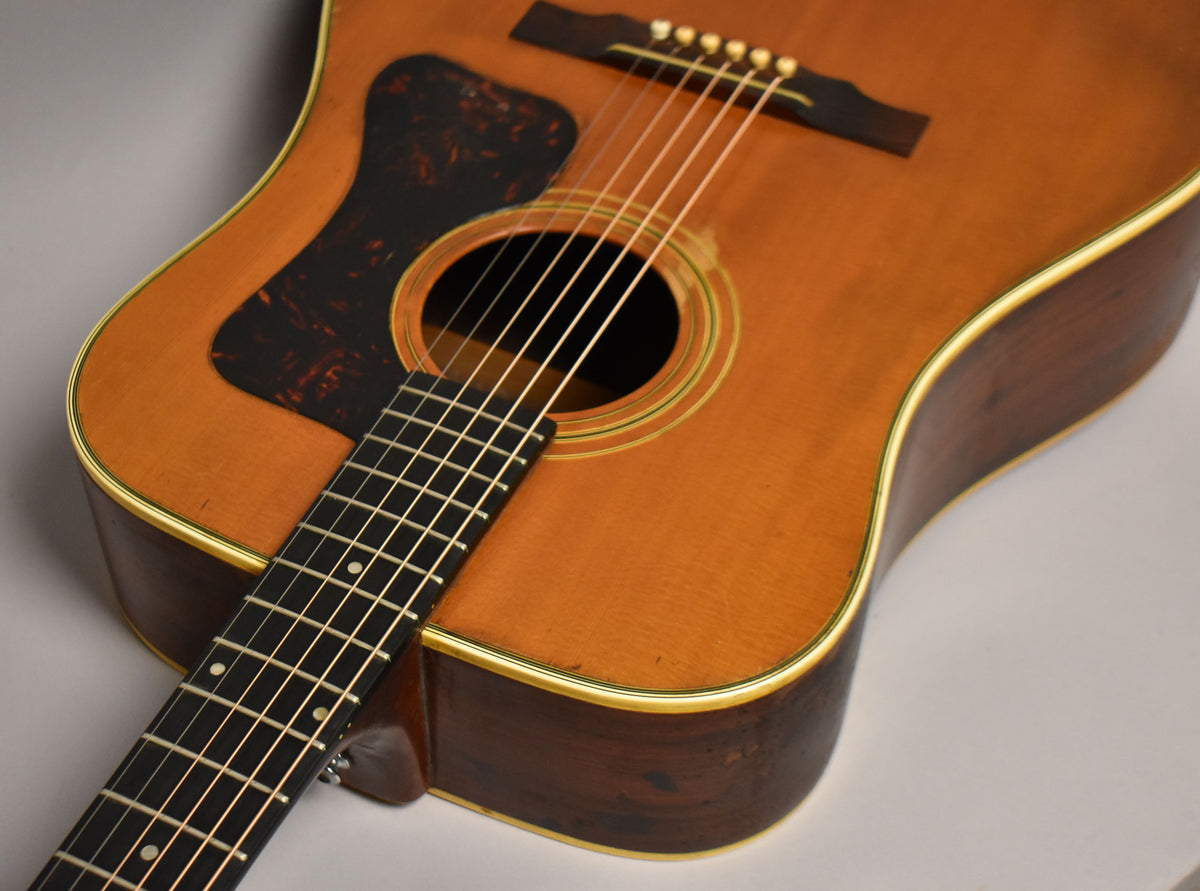 Close-up of the natural-finish acoustic guitar's body, showing the dark pickguard, soundhole, bridge, and lower fretboard.