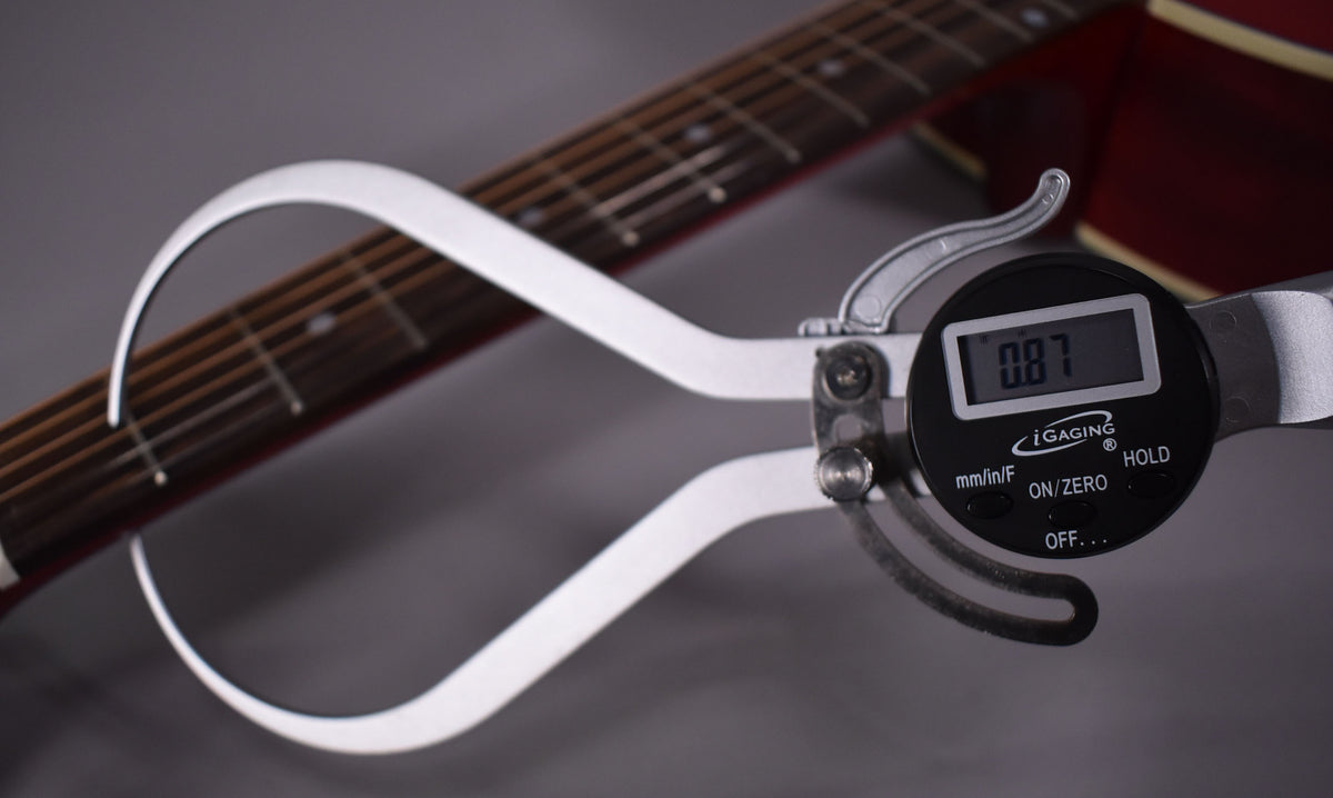 A digital caliper measuring the action of the guitar strings above the fretboard, displaying "0.87" on its screen.