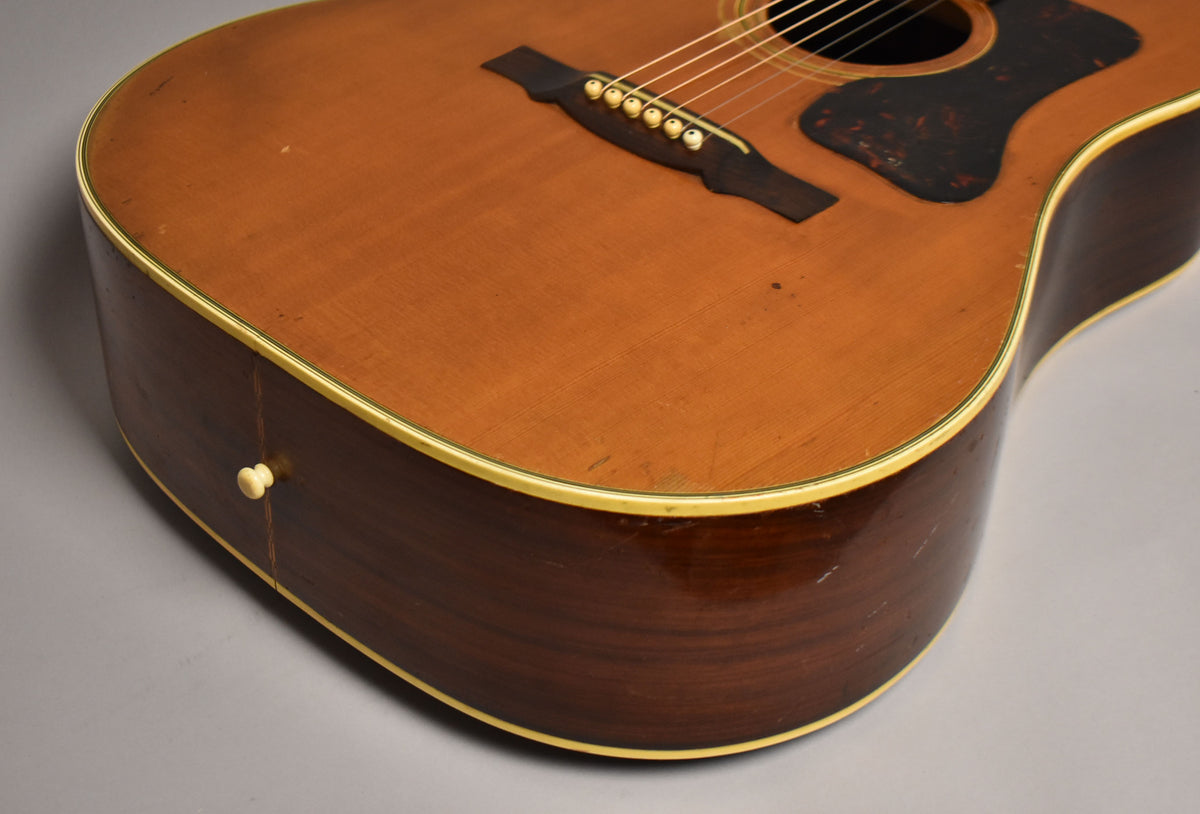 The lower bout of a natural acoustic guitar, showing the dark brown sides, cream binding, and a cream end pin.