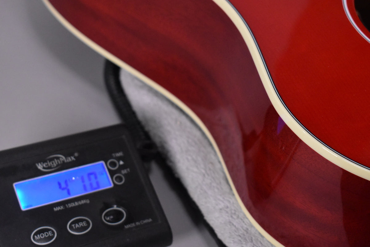 The guitar's cherry red body, with cream binding, is shown resting on a soft case, next to a digital scale displaying "4.10".