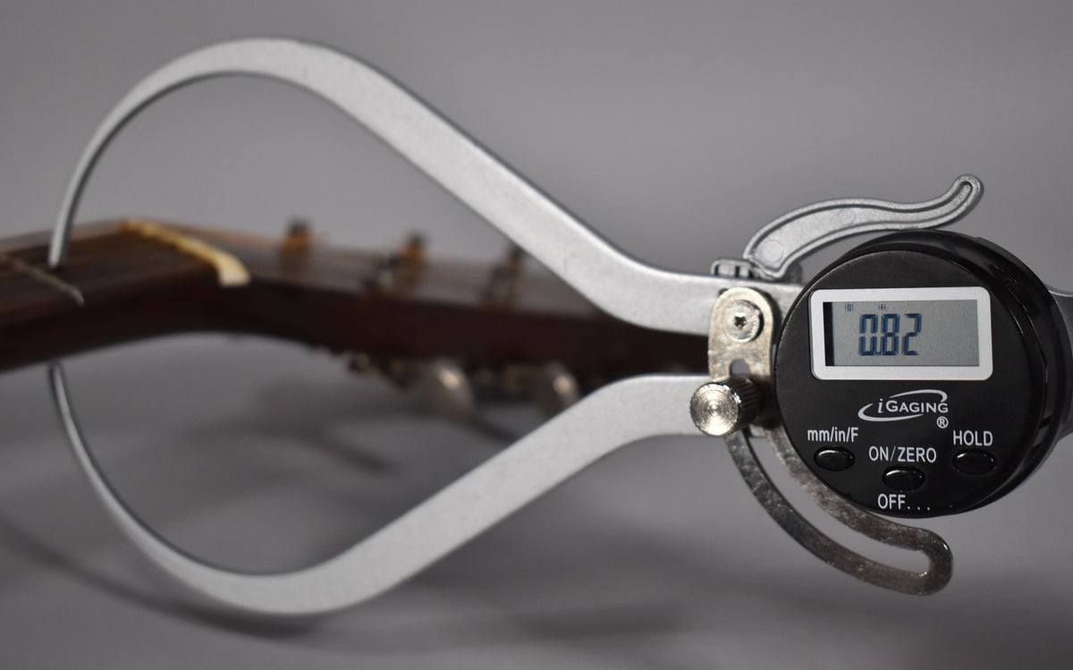 A digital caliper measures the guitar's nut, showing 0.82, with the dark brown headstock and tuning pegs blurred in the background.