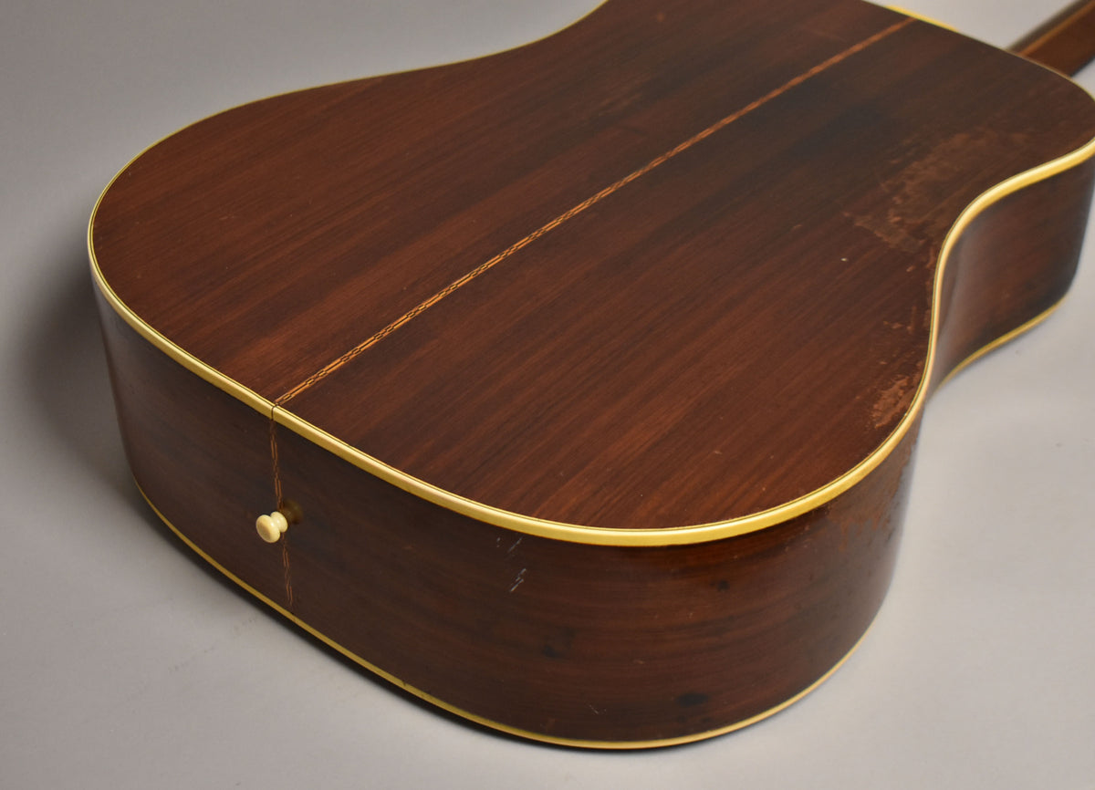 The lower bout of a dark brown acoustic guitar, showing a cream end pin, binding, and significant finish wear on the back.