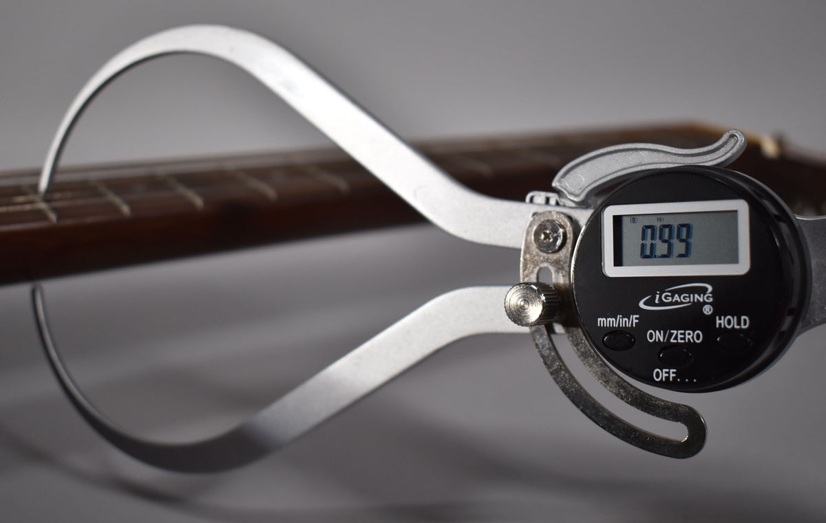 A digital caliper measures the guitar's dark brown fretboard, showing a reading of 0.99 on its black display.