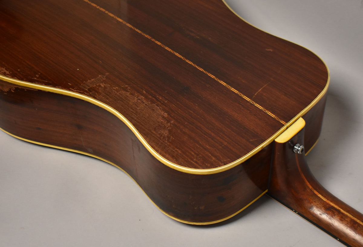 The guitar's dark brown back and side, with a light binding, show significant finish wear and scratches, especially on the lower bout.