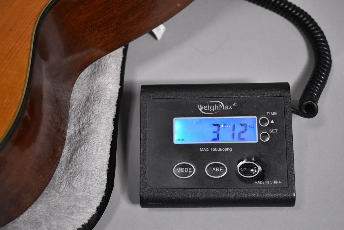 The guitar's dark brown neck heel and lower bout are visible, resting in a plush grey case next to a digital scale reading "3.12".