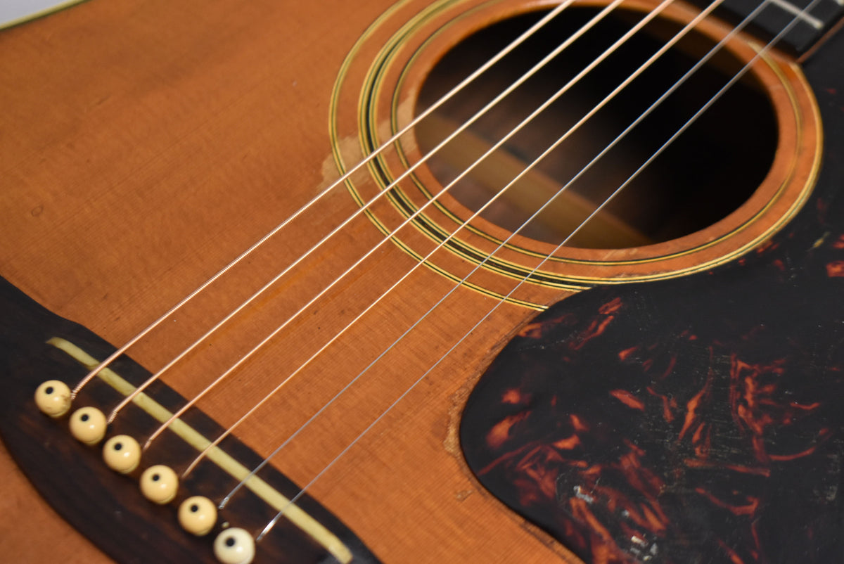 Close-up of the natural-finish acoustic guitar's body, showing the soundhole, tortoise pickguard, bridge with six cream pins, and string wear.