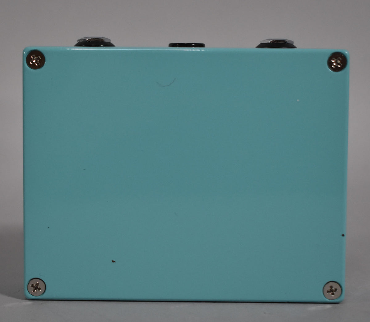 The turquoise pedal is viewed from the back, showing four silver screws and two black knobs along the top edge.