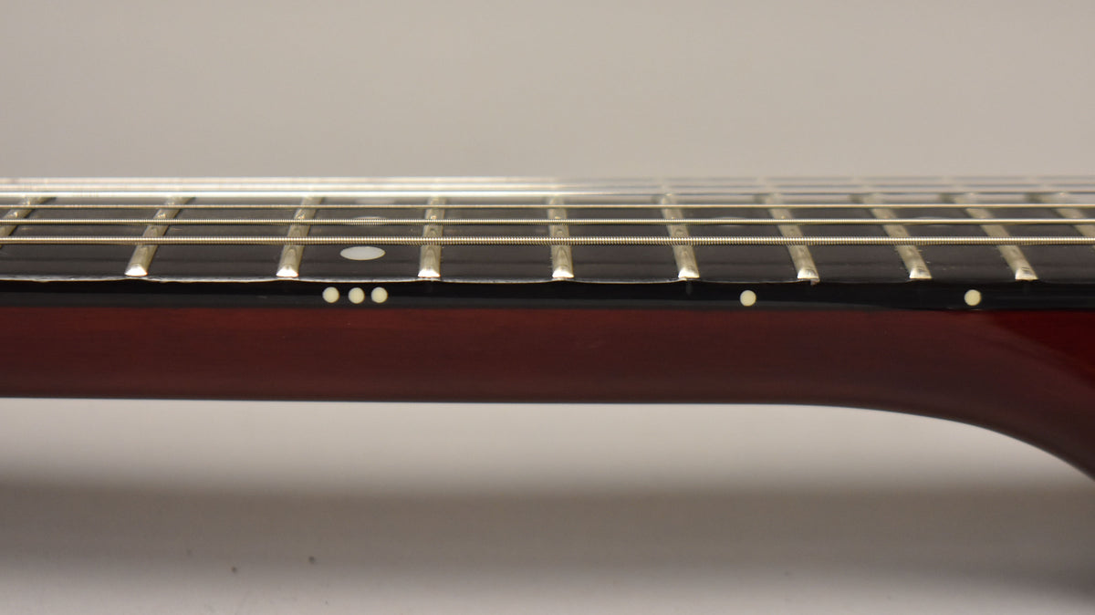 The cherry red guitar neck, with a dark fretboard and white dot inlays, is viewed from the side, showing frets and strings.