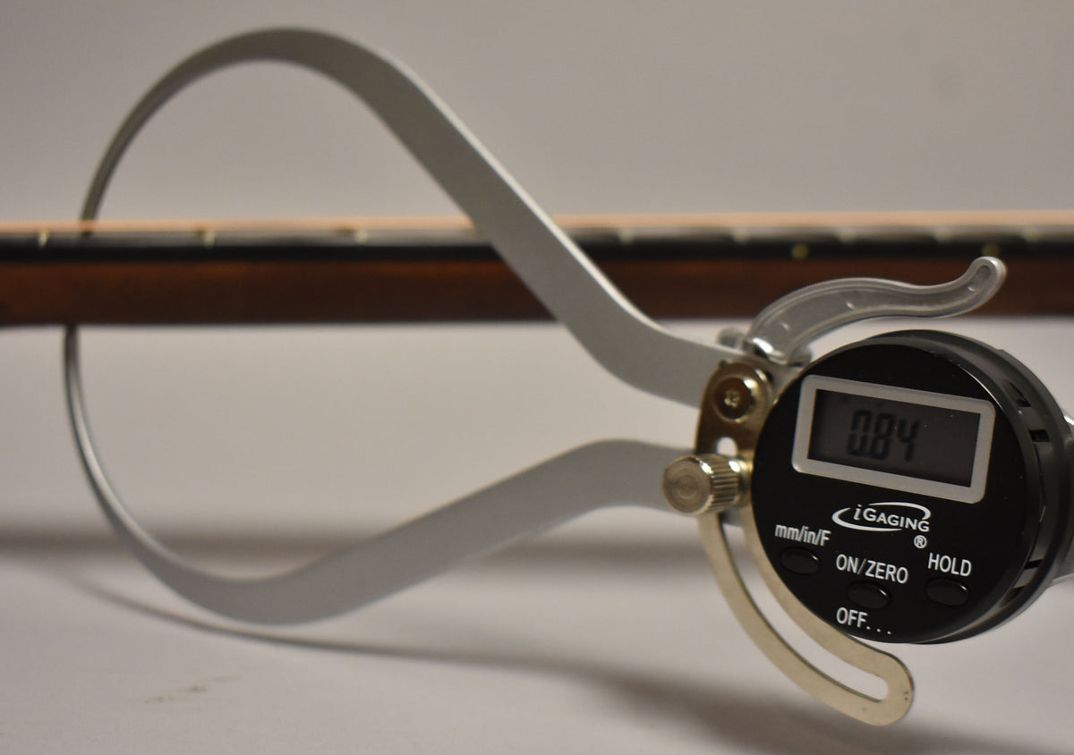 A digital caliper measures the guitar's neck thickness, showing "0.84" on its screen, with the dark brown neck in the background.