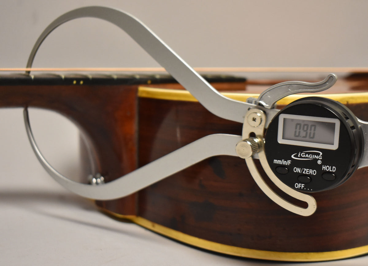 A digital caliper measures the string height at the 12th fret of the natural finish acoustic guitar, showing 0.90.