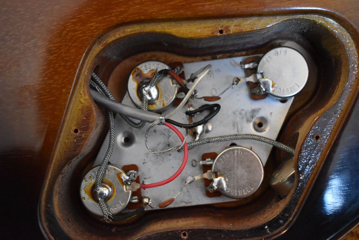 Close-up of the guitar's control cavity, revealing four silver potentiometers, wiring, and a metal plate against the dark wood.