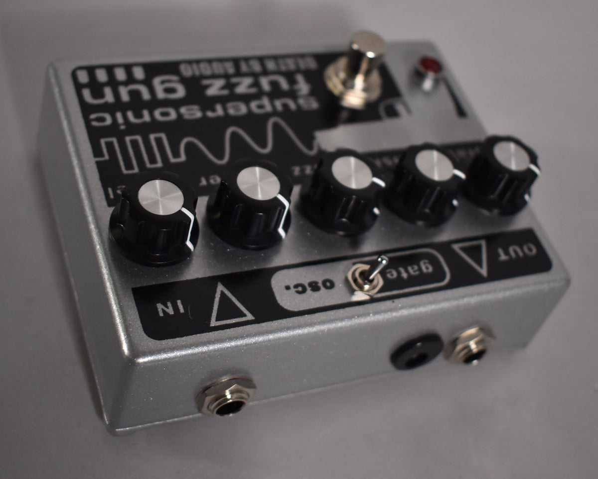 Silver fuzz pedal, angled from the left, showing five black knobs, a toggle switch, and input/output jacks.