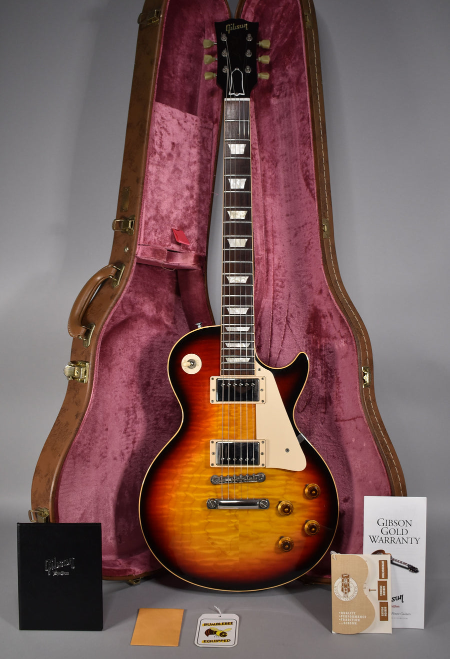 Full-length view of a glossy tri-burst Les Paul with cream binding, pickguard, and pickup rings, standing in its open brown case.