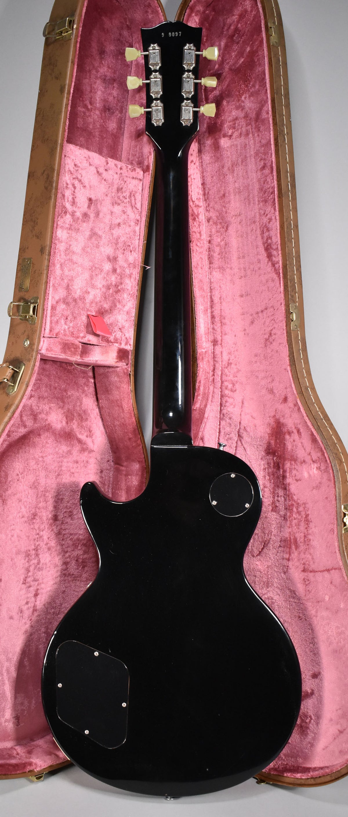 The guitar's glossy black back and neck are visible, with six cream tuning pegs on the headstock, resting in a pink velvet case.