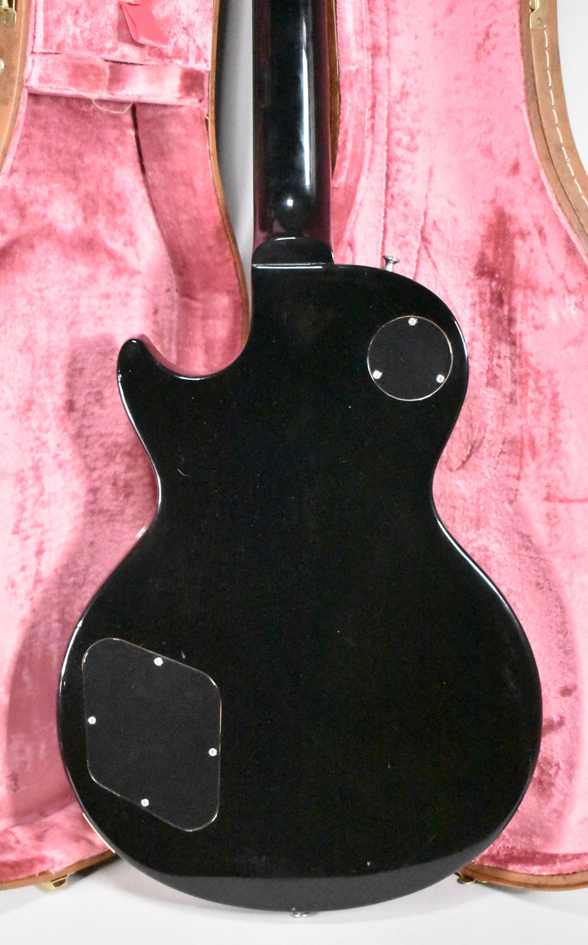 The guitar's glossy black back and neck are visible, with two control cavity covers, against a pink plush-lined case.