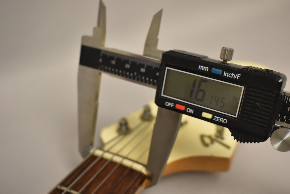 A digital caliper measures the guitar's nut width at 1.6 inches, with the Olympic White headstock and dark fretboard visible.