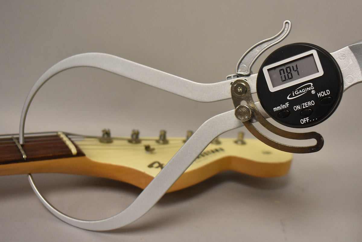 A digital caliper measures the guitar's neck thickness at the first fret, showing 0.84 inches, with the headstock visible.