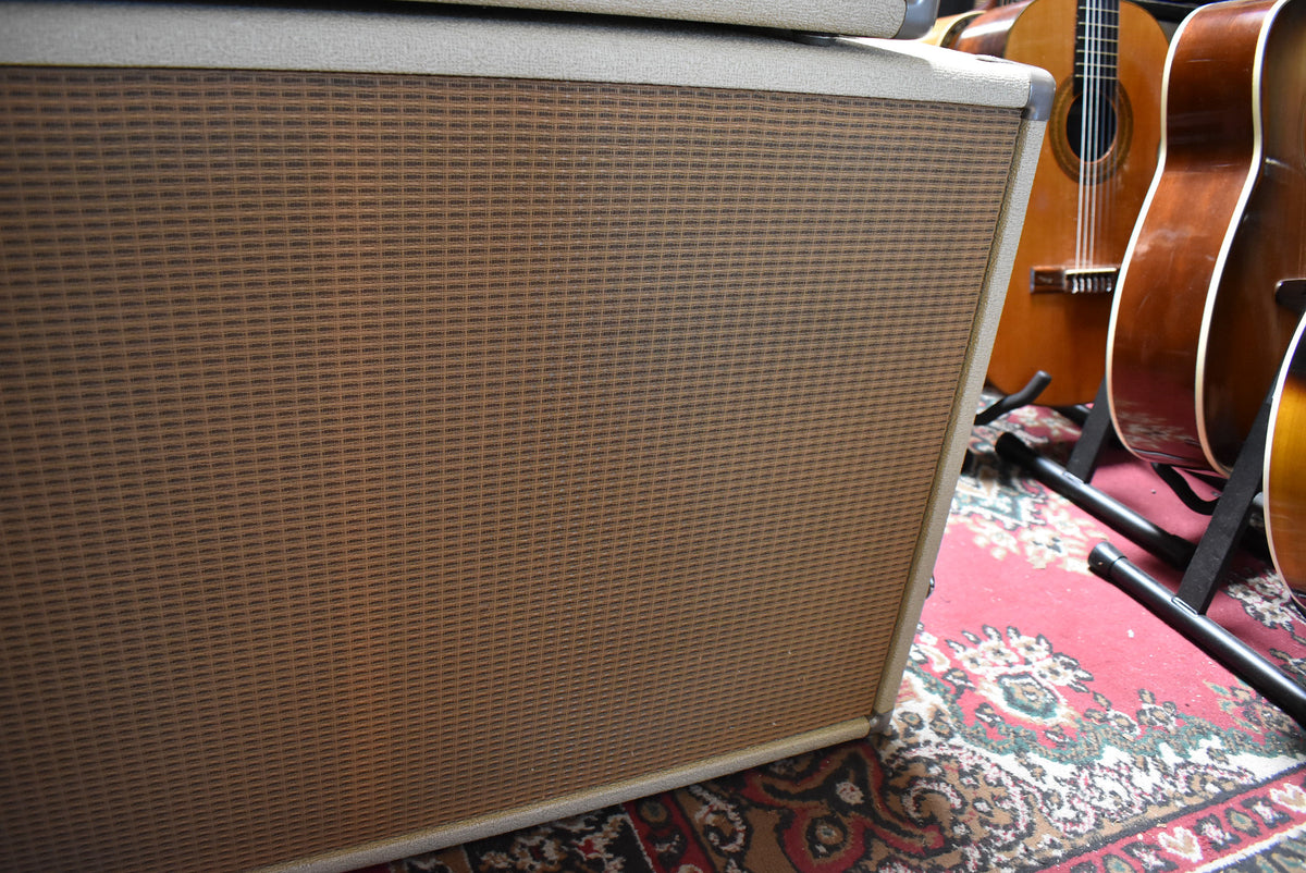 The blond 2x12 cabinet is angled, showing its light brown grille cloth with a dark brown grid pattern, and some dark discoloration on the fabric.