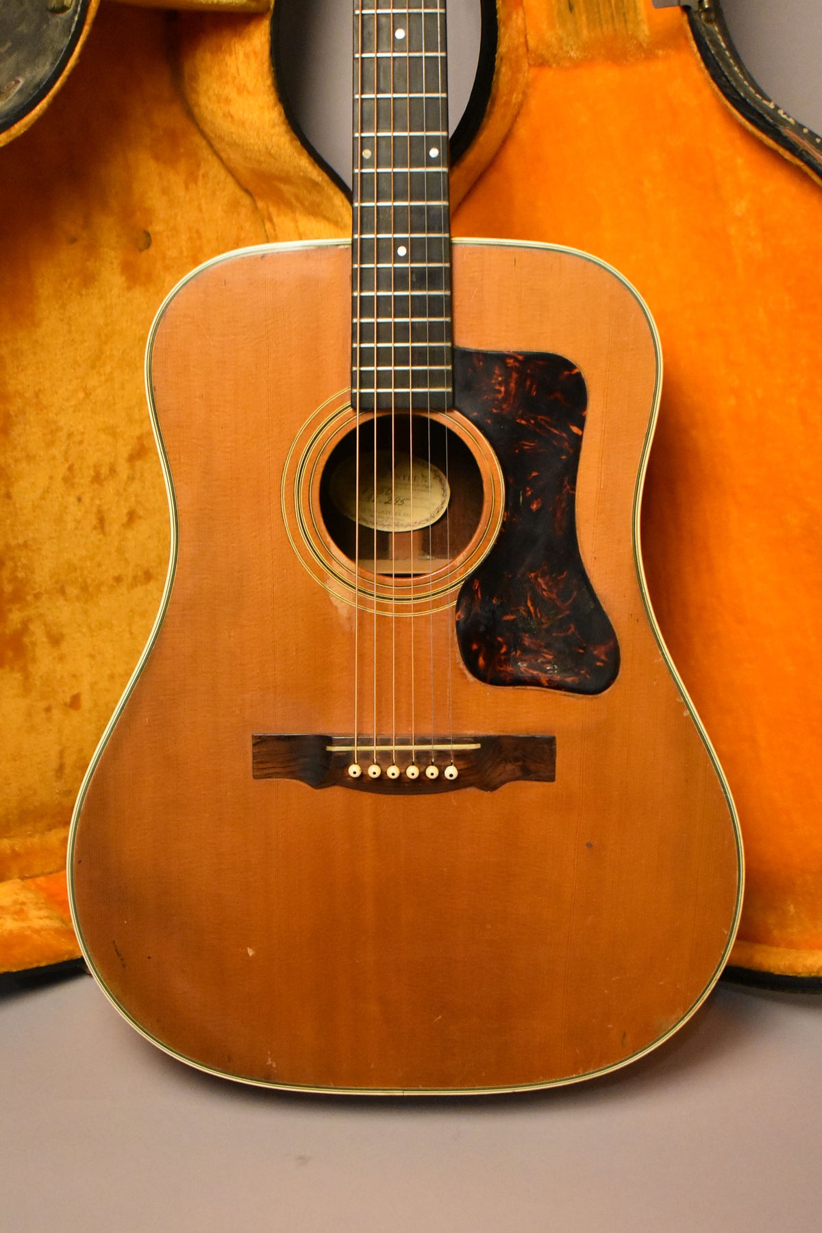Full front view of a natural finish acoustic guitar with a tortoiseshell pickguard, showing wear and checking on the body.