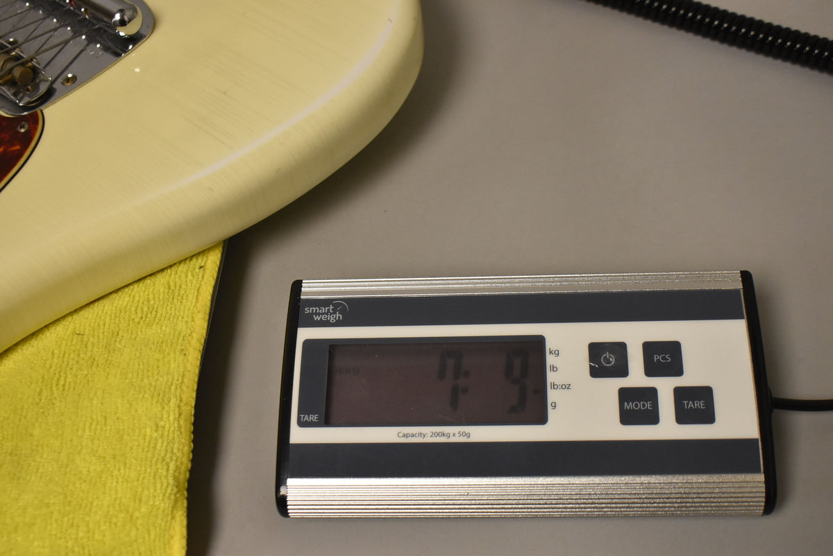 The guitar's Olympic White body, with visible nitro checking, rests on a yellow cloth next to a digital scale displaying "7.9" lbs.
