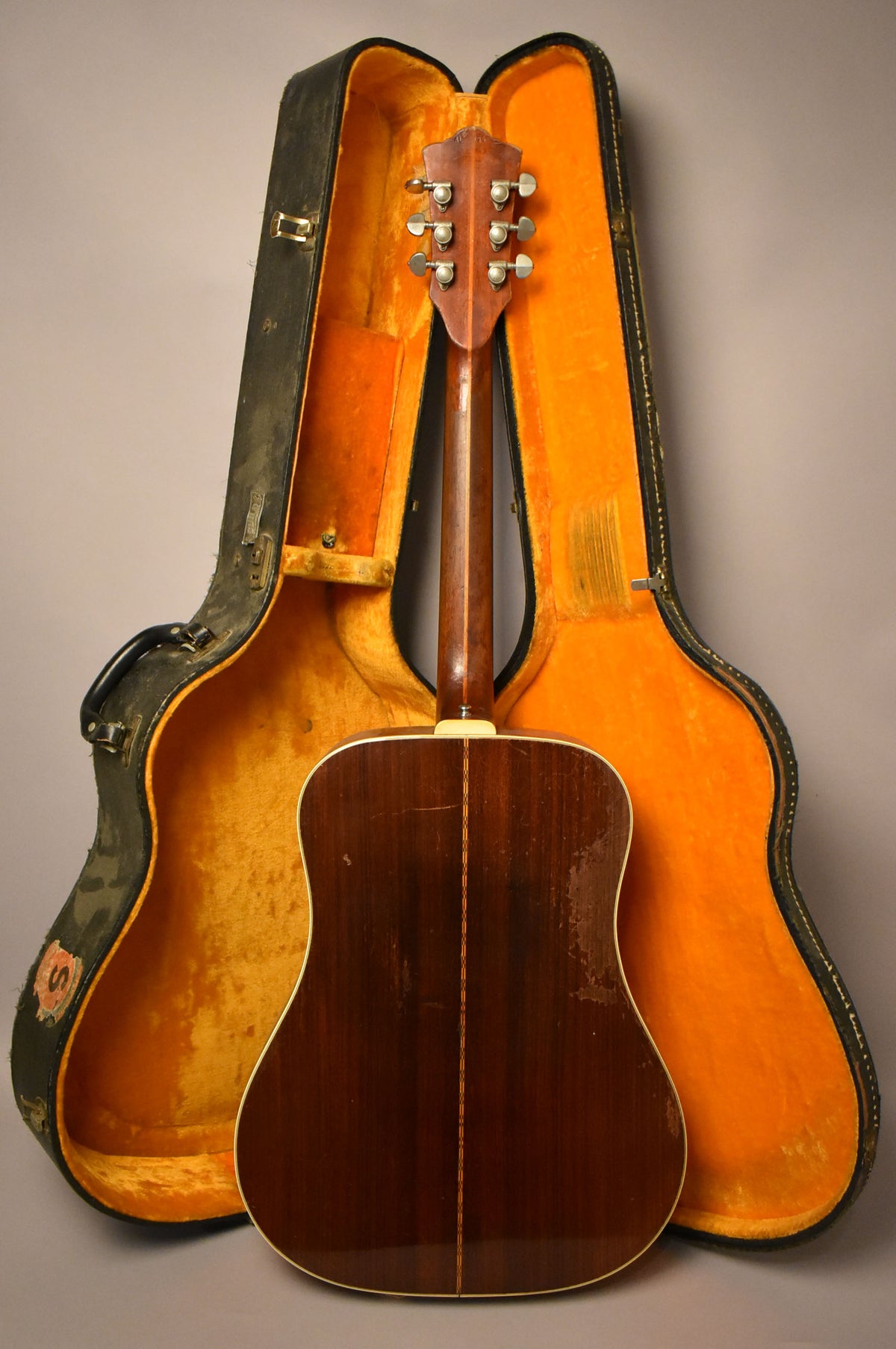 The guitar's dark wood back and neck face away, showing six tuners and heavy finish wear, with its open case behind.