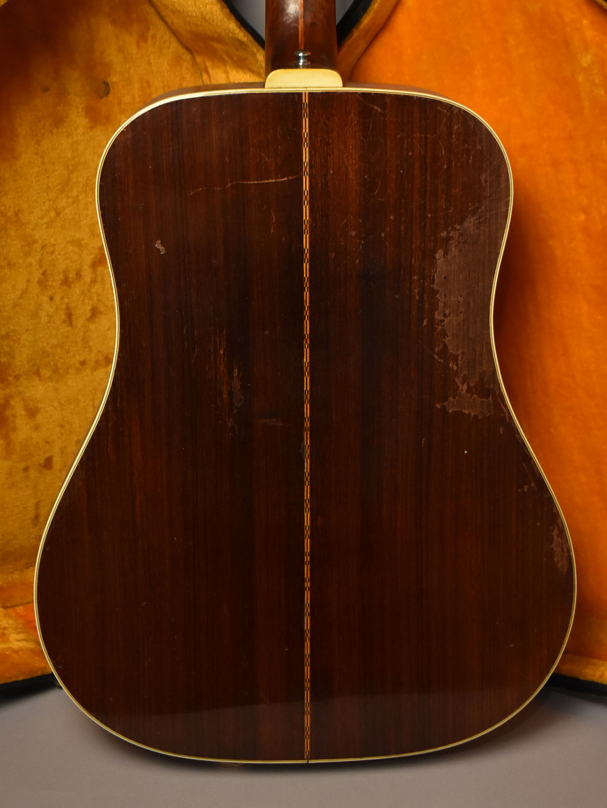 The guitar's dark brown back shows a central light wood stripe, binding, and significant finish wear and scratches, with an orange case lining visible.