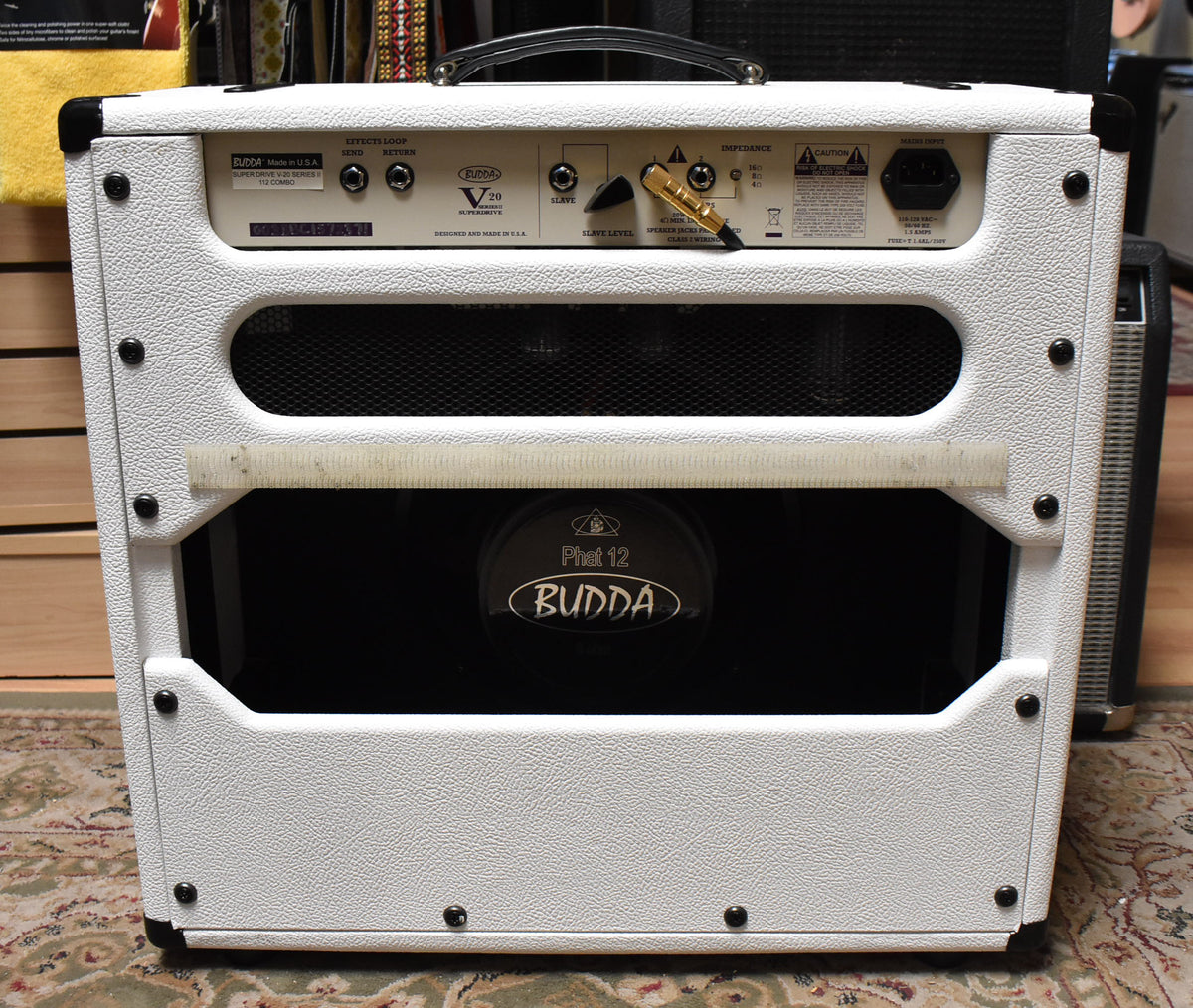 The white textured combo amp is seen from the rear, revealing a black speaker, a gold-tipped switch, and various input jacks.