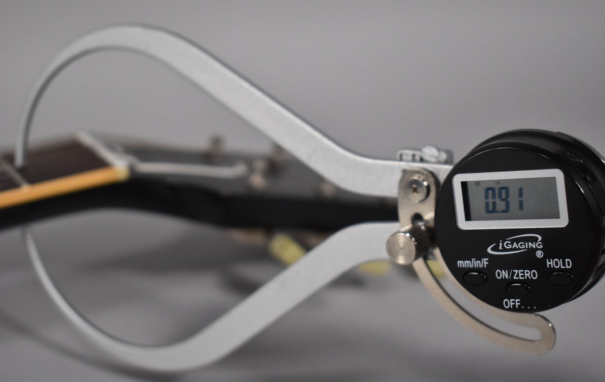 A digital caliper measures the guitar's nut width, displaying "0.91" on its screen, with the dark headstock blurred in the background.