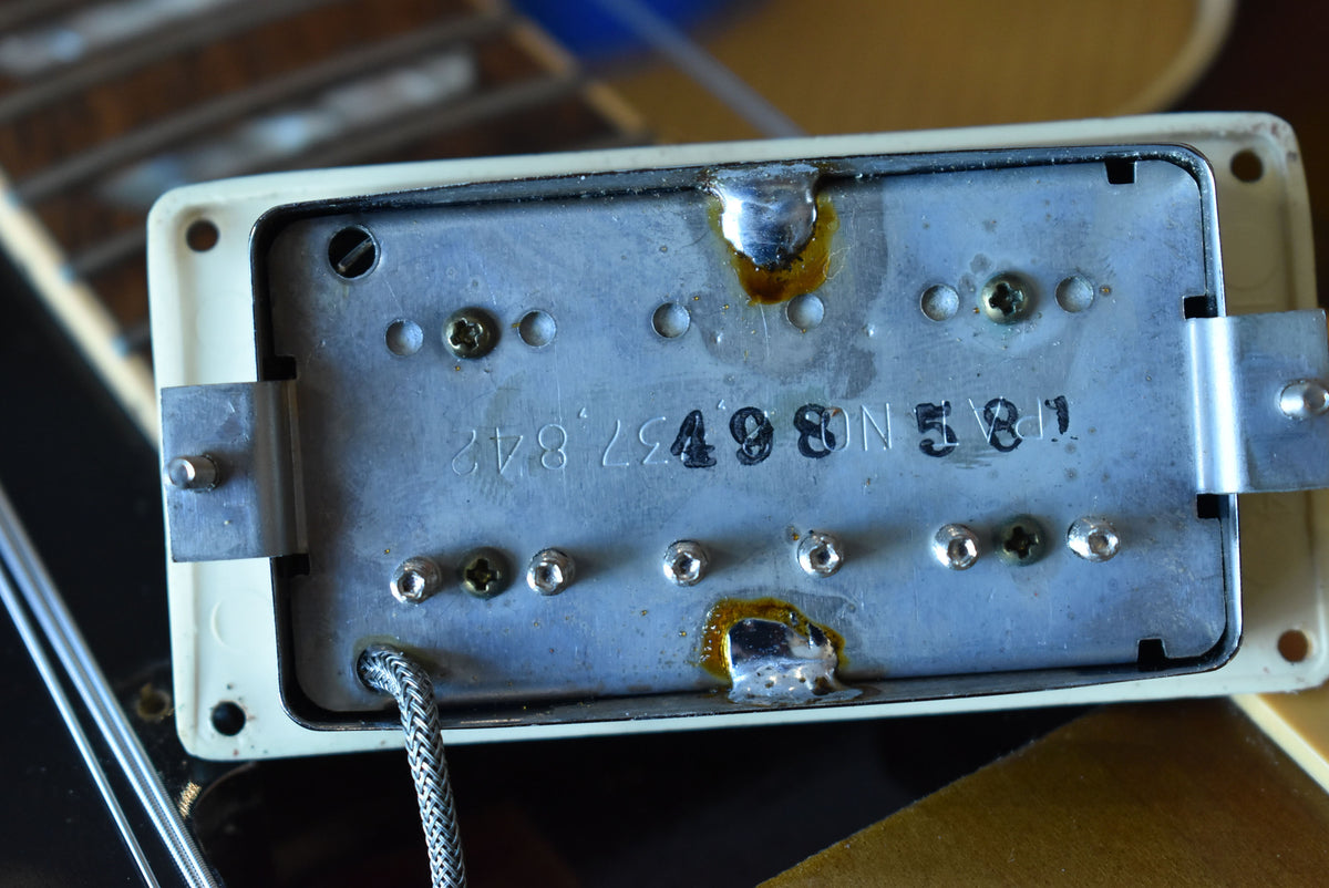 Close-up of the underside of a cream humbucker pickup, showing the metal baseplate with stamped numbers and solder joints.