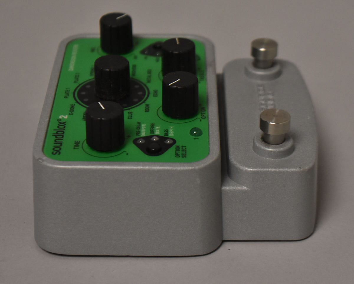 The gray pedal, with a green top panel and black knobs, is angled right, showing two silver footswitches on its side.