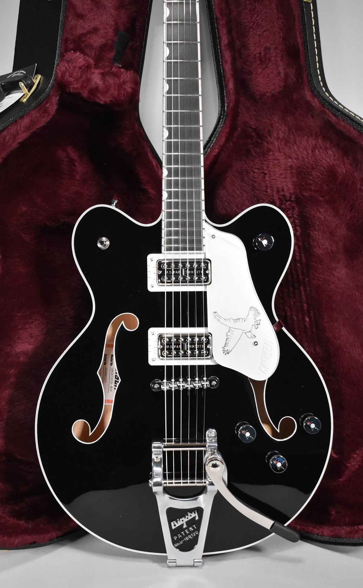 A black hollow-body electric guitar with white binding and pickguard, chrome hardware, and a Bigsby vibrato, in a red plush case.